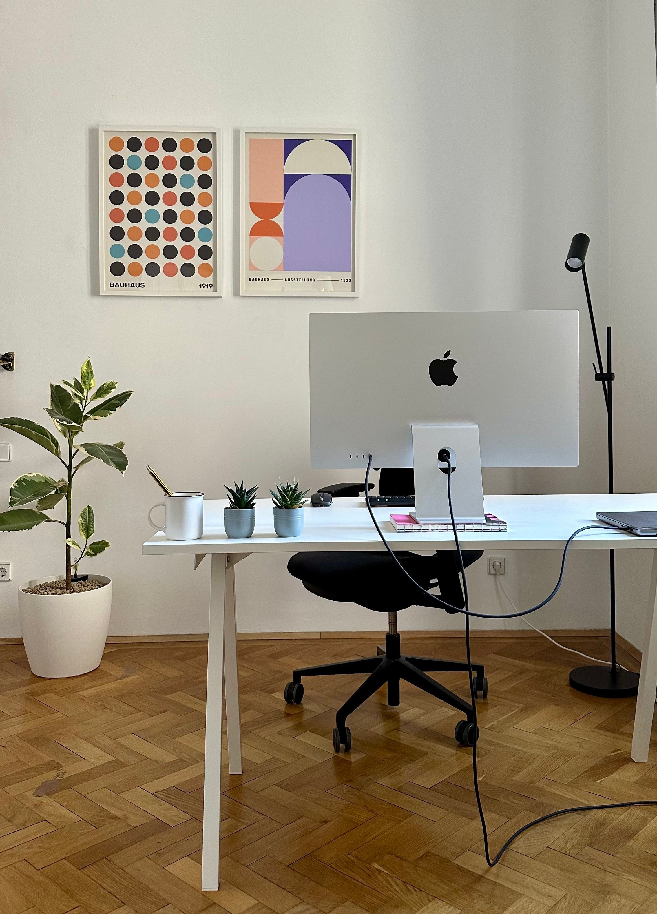 Minimalist home office with a white desk and an Apple desktop computer centered on top. A black office chair sits beneath the desk, and a black floor lamp stands to the right. On the desk are small potted plants, a white mug with a spoon, notebooks, and a closed laptop. Two framed abstract Bauhaus-style prints hang on the white wall above. A tall leafy plant in a white pot stands to the left, and the floor is light wood in a herringbone pattern.